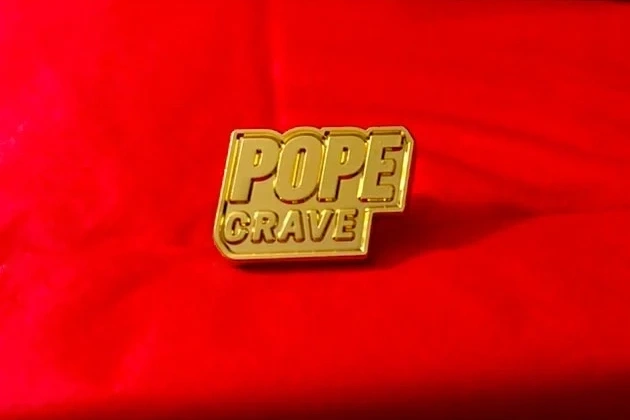 Gold Popecrave Pin laying on top of a red wrapped Conclave Zine.